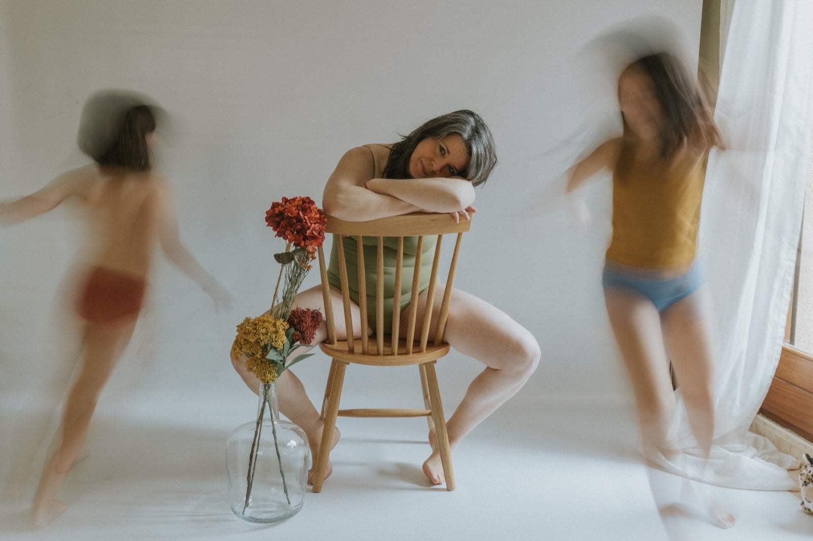 Cristina Pujol (Ojos abiertos) sentada en una silla, flores en florero a su lado. Sus hijas bailando alrededor. Foto de Cristina Pujol